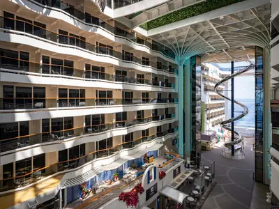 Vogelperspektive auf die World Promenade der MSC World America mit der spiralförmigen Trockenrutsche und vielen Balkonkabinen. Im Hintergrund ist das Meer sichtbar. Ein lebhaftes, modernes Bordleben.