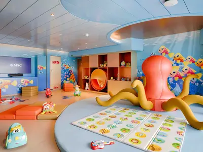 Ein lebhafter Kinderbereich an Bord der MSC World America mit einem riesigen orangefarbenen Kraken, Fisch-Wandmalereien, einem TV-Bildschirm und Spielzeugen wie einem roten Roller und weichen Matten zum Spielen.