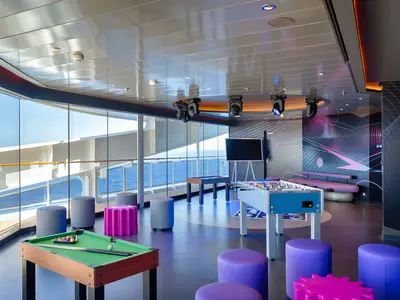 Heller und moderner Teen Club an Bord der MSC World America mit Billardtisch, Kicker und lila Hockern, großen Fenstern und Blick auf das Meer.