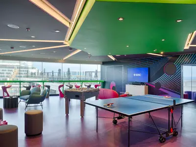Ein lebhafter Young Club an Bord der MSC World America mit einem Tischfußballtisch, einer Tischtennisplatte und modernen Sitzgelegenheiten in verschiedenen Farben. Große Fenster bieten Meerblick, während an einer Wand ein Fernseher mit dem MSC Logo hängt. Das Interieur ist farbenfroh und modern gestaltet.
