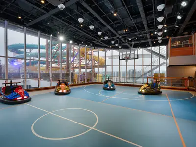 Ein Indoor-Sportplex an Bord der MSC World America. Eine hellblaue Spielfläche mit Basketballkorb und vier bunten Bumper Cars. Grosse Fenster geben den Blick auf farbige Wasserrutschen frei.