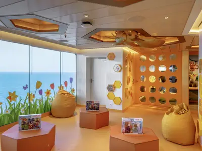 Ein heller, orangefarbener Kinderbereich an Bord der MSC World America mit großen Fenstern zum Meer, verspielten Blumenstickern, Sitzsäcken und Bienenstock-Dekorationen. Auf den Tischen liegen Puzzles.