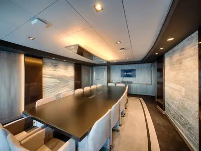 Eleganter Konferenzraum an Bord der MSC World America. Langer dunkler Tisch, viele helle Stühle. Moderne Wände, Kunst, Bildschirm mit MSC Logo und warme Beleuchtung.