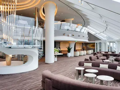 Helles und modernes Lounge-Interieur der MSC World America. Eine geschwungene Glas-Stahltreppe, viele lila Sofas und eine obere Restaurantebene sind sichtbar.