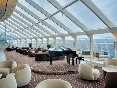 Blick in die helle und moderne Top Sail Lounge der MSC World America mit großen Fenstern zum Meer, bequemen Sofas, Sesseln und einem schwarzen Flügel im Zentrum. Die Decke ist aus Glas.