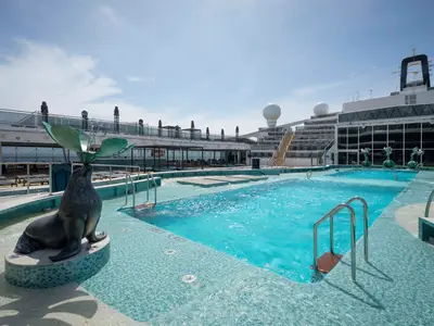 Ein sonniger Poolbereich an Bord der MSC World America mit türkisfarbenem Wasser, Liegen, einer Seelöwen-Skulptur und modernen Glasfassaden. Das Deck ist in hellen Blautönen gehalten.