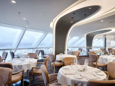 Elegantes Restaurant an Bord der MSC World America mit Tischen, Stühlen und grossen Fenstern, die den Blick auf das Meer freigeben. Die Decke ist mit geschwungenen Lichtinstallationen verziert.