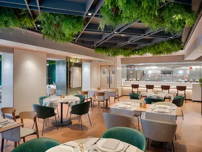 Ein stilvolles Spezialitätenrestaurant an Bord der MSC World America. Der Raum ist mit modernen Tischen, grünen Stühlen und einer offenen Küche mit Kupfertöpfen ausgestattet. Deckenbegrünung sorgt für eine frische Atmosphäre.
