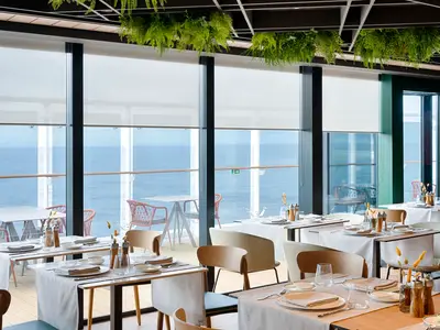 Helles Restaurant an Bord der MSC World America. Elegant gedeckte Tische und Stühle, grosse Fenster mit Meerblick. Hängepflanzen verzieren die Decke.