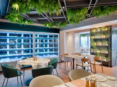 Das Eataly Restaurant an Bord der MSC World America zeigt moderne Tische, grüne Stühle und beleuchtete vertikale Gärten mit frischen Pflanzen. Üppiges Grün hängt von der Decke.