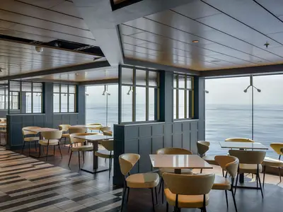 Restaurant an Bord der MSC World America. Helle Tische, cremefarbene Stühle und große Fenster bieten weiten Meerblick. Dunkle Paneele mit goldenen Akzenten und gemusterter Boden.