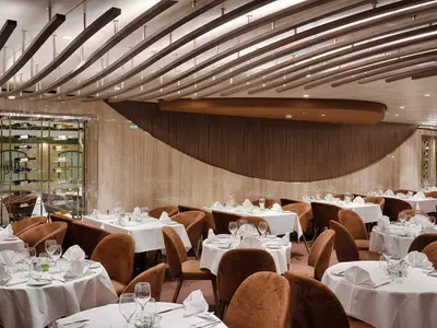 Blick in das moderne Restaurant Les Dunes an Bord der MSC World America. Viele Tische mit weißen Tischdecken und braunen Stühlen sind gedeckt. Eine Weinwand aus Glas ist sichtbar, die Wände sind mit Holz verkleidet.