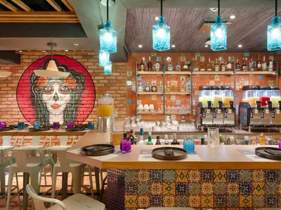 Blick in das farbenfrohe Spezialitätenrestaurant Hola! Tacos & Cantina an Bord der MSC World America mit einer Bar, bunten Fliesen und Wandkunst. Auf dem Bartresen stehen Getränkespender und Gläser bereit.