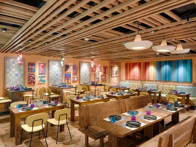 Das farbenfrohe "Hola! Tacos & Cantina" Spezialitätenrestaurant an Bord der MSC World America zeigt Holztische, Stühle und Bänke. Die Wände sind mit mexikanischen Fliesen und Kunstwerken dekoriert. Eine Holzlamellendecke und besondere Deckenleuchten runden das Ambiente ab.