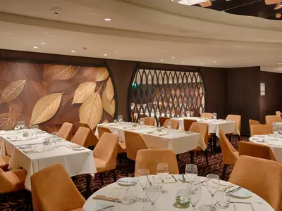 Blick in das La Foglia Restaurant an Bord der MSC World America mit gedeckten Tischen, beigen Stühlen und einer Wanddekoration aus Blättern.