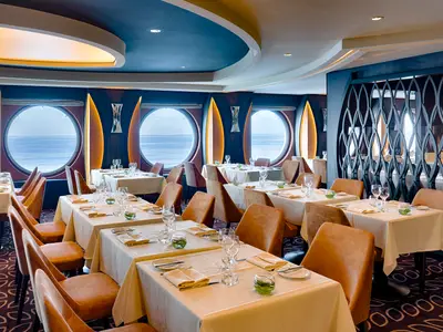 Ein elegantes Restaurant an Bord der MSC World America mit mehreren Tischen, die mit weissen Tischdecken und Besteck gedeckt sind. Grosse runde Bullaugen bieten Meerblick.