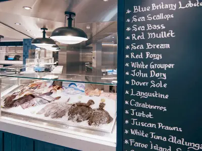 Frisches Seafood auf Eis im Spezialitaetenrestaurant der MSC World America. Tafel listet handgeschriebene Gerichte: Blue Brittany Lobster, John Dory. Heller, moderner Innenraum.