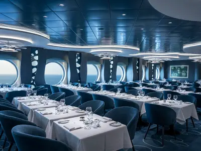 Elegantes Restaurant an Bord der MSC World America. Zahlreiche weiß gedeckte Tische, blaue Stühle und runde Fenster mit Meerblick. Moderne Einrichtung.