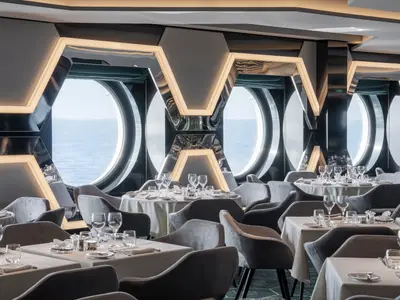 Ein modernes Restaurant an Bord der MSC World America mit elegant gedeckten Tischen und bequemen Samtstühlen. Große runde Bullaugen bieten einen weiten Blick auf das offene Meer. Die Wände sind mit markanten, beleuchteten Hexagon-Elementen verziert, die dem Raum eine futuristische Atmosphäre verleihen.