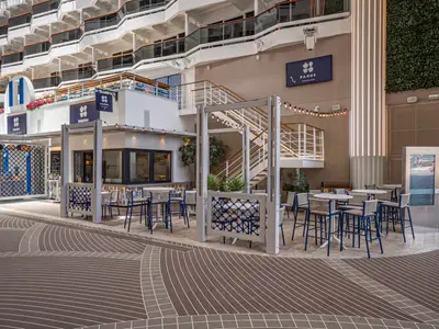 Blick auf den Paxos Greek Taverna Bereich an Bord der MSC World America mit blau-weißen Dekorationen, Tischen, Stühlen und Pflanzen unter hellen Lichtern, eingebettet in einer mehrstöckigen Promenade.