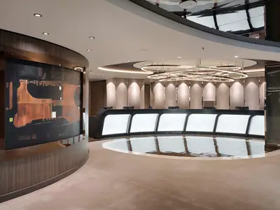 Eine elegante, geschwungene Rezeption an Bord der MSC World America mit beleuchteten Schaltern, einem großen abstrakten Kunstwerk und einer modernen Lichtinstallation an der Decke. Der Boden reflektiert Teile der Decke.