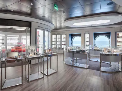 Innenansicht der modernen Shopping Gallery an Bord der MSC World America. Mehrere beleuchtete Vitrinen präsentieren Uhren und Sonnenbrillen. Zwei runde Bullaugen bieten Meerblick.