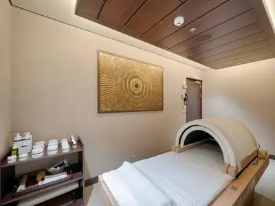 Ein heller, moderner Spa-Behandlungsraum an Bord der MSC World America. Eine weiße Liege mit einem bogenförmigen Infrarot-Gerät steht im Vordergrund, daneben ein Regal mit Pflegeprodukten. Eine goldene Wandkunst schmückt die Wand.