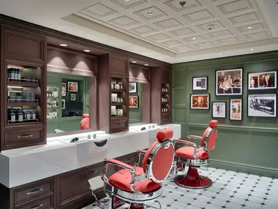 Eleganter Herrenfriseur an Bord der MSC World America. Rote Friseurstühle, weiße Marmor-Waschtische und grüne Holzpaneele mit Bildern. Braune Regale präsentieren Pflegeprodukte.