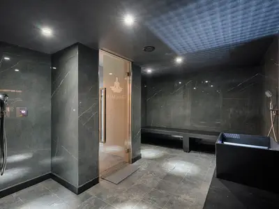 Moderner Dampfbadbereich an Bord der MSC World America. Dunkle Marmorwände, eine Glastür mit Meditationssymbol. Im Raum sind eine lange Sitzbank, ein schwarzes Becken und elegante Duscharmaturen zu erkennen.