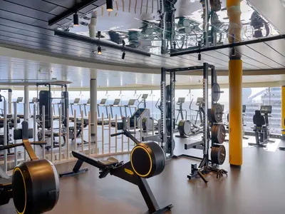 Ein modernes Fitnessstudio an Bord der MSC World America. Man sieht Rudergeräte, Hantelstangen und Laufbänder vor großen Fenstern mit Blick auf das offene Meer.