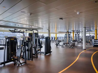 Helles Fitnessstudio an Bord der MSC World America, ausgestattet mit vielen Technogym-Geräten. Große Fenster bieten Meerblick, der Boden ist dunkel mit gelben Laufbahn-Linien. Keine Personen sichtbar.