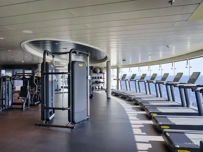 Ein helles, modernes Fitnessstudio an Bord der MSC World America. Eine Reihe von Laufbändern steht vor großen Panoramafenstern mit Blick auf das blaue Meer. Im Vordergrund befinden sich schwarze Krafttrainingsgeräte und Hantelablagen. Der Raum ist leer von Personen.