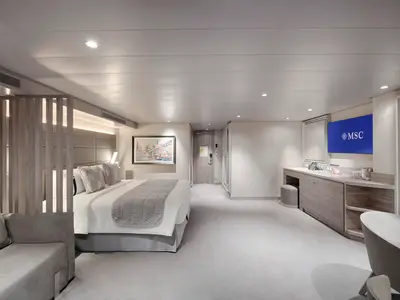 Geräumige Grand Suite Aurea an Bord der MSC World America. Es zeigt Doppelbett, Wohnbereich mit Sofa, Schreibtisch und TV, getrennt durch eine Holzlamellenwand. Modernes Design.