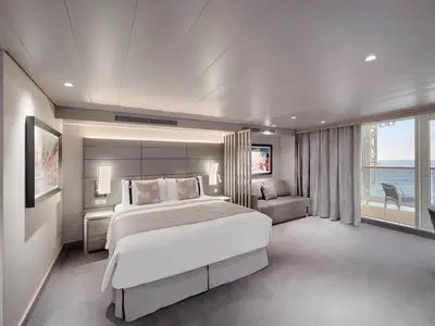 Blick in eine geräumige, moderne Kabine der MSC World America mit großem Doppelbett, Sofa und einem Balkon, der eine weite Aussicht auf das offene Meer bietet. Die Einrichtung ist in Grau- und Cremetönen gehalten, mit eleganten Akzenten und indirekter Beleuchtung.