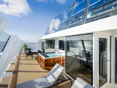 Private Terrasse einer Owner's Suite an Bord der MSC World America. Ein Whirlpool, eine Sonnenliege und ein Esstisch sind zu sehen. Im Hintergrund spiegelt sich das Meer in den Glasfassaden. Blauer Himmel.