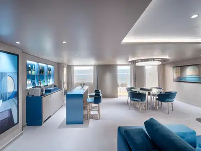 Eine geräumige, moderne Owners Suite an Bord der MSC World America mit blauem Dekor, einer stilvollen Bar, einem Esstisch und großen Fenstern zum Meer.