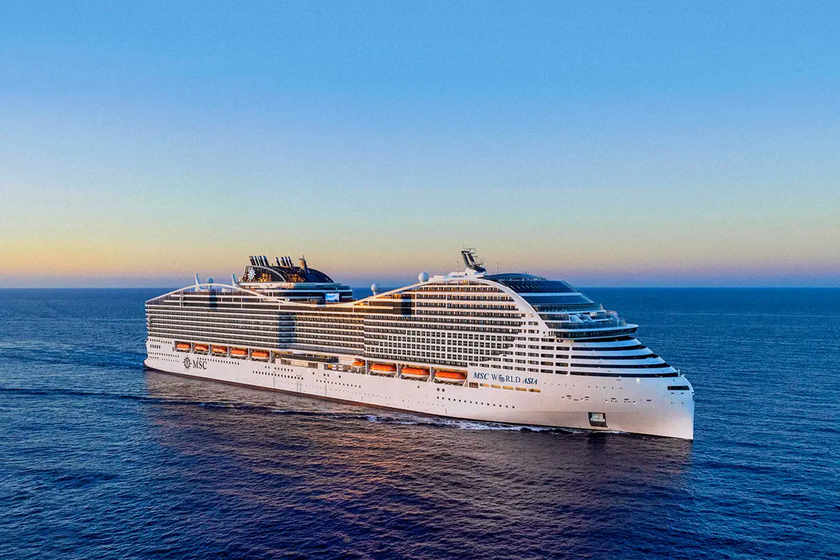 Das moderne Kreuzfahrtschiff MSC World Asia fährt auf offenem Meer. Der Himmel zeigt Blau- und Orangetöne des Sonnenuntergangs. Das weiße Schiff ist in Seitenansicht mit vielen Decks und Rettungsbooten zu sehen.