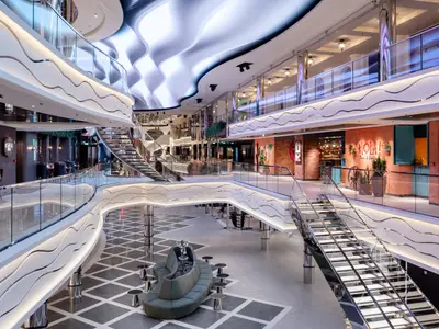 Weitwinkelansicht des mehrstöckigen, eleganten Atriums an Bord der MSC World Asia, mit wellenförmigen Geländern, hell erleuchteten Decken und einer Auswahl an Restaurants, darunter HOLÀ! Tacos & Cantina.