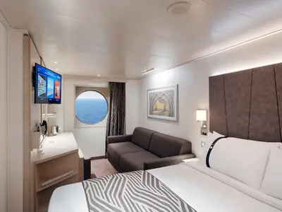 Helle Aussenkabine an Bord der MSC World Asia. Man sieht ein Doppelbett, ein Sofa, einen Schreibtisch mit TV und ein Bullaugenfenster mit Blick auf das offene Meer.