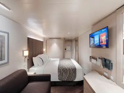 Moderne Aussenkabine an Bord der MSC World Asia mit Doppelbett, Sofa, Flachbild-TV und Schreibtisch. Helle Hölzer und neutrale Farben dominieren das Design.