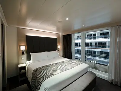 Innenansicht einer stilvollen Balkonkabine an Bord der MSC World Asia. Im Vordergrund ein gemuetliches Doppelbett mit Zebra-Decke und Sitzbank. Durch die Balkontuer sind weitere Schiffsbalkone der MSC World Asia zu sehen. Eine weisse Gardine haengt am Fenster.