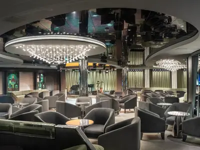 Eine stilvolle Bar-Lounge an Bord der MSC World Asia, mit bequemen Polstersesseln, kleinen Tischen und einer Bühne mit Klavier und Schlagzeug. Die Decke ist verspiegelt, ein grosser runder Kronleuchter mit vielen Leuchten hängt mittig. Elegante Vorhänge und moderne Kunstbilder schmücken den Raum.