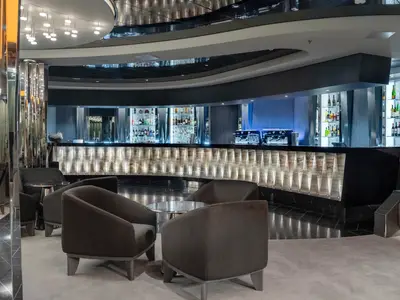 Die stilvolle Dolce Vita Bar an Bord der MSC World Asia präsentiert eine lange, beleuchtete Bartheke mit Getränken und Flaschen. Dunkle Sessel und runde Tische laden zum Verweilen ein.