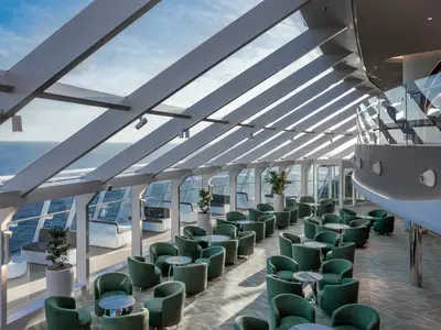 Eine luxuriöse Bar-Lounge mit bodentiefen Fenstern und schrägem Glasdach bietet weitreichenden Meerblick an Bord der MSC World Asia. Grüne Sessel und kleine Tische laden zum Verweilen ein.