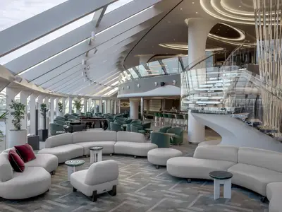 Elegante Top Sail Lounge an Bord der MSC World Asia. Der helle, weitläufige Raum bietet weisse Sofas, grüne Sessel, eine Bar und eine moderne Glas-Treppe.