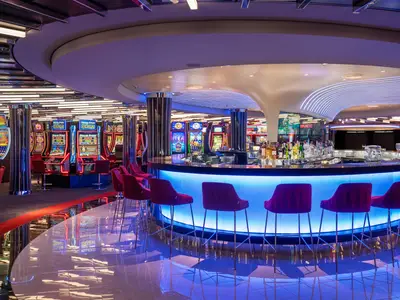Blick in das moderne Casino an Bord der MSC World Asia. Eine blau beleuchtete Bar mit violetten Barhockern im Vordergrund, dahinter zahlreiche leuchtende Spielautomaten.