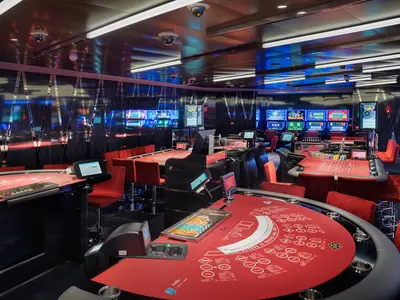 Modernes Casino an Bord der MSC World Asia. Rote Spieltische, Chips und Stühle im Vordergrund. Dahinter leuchten Reihen von Spielautomaten. Dunkle, glänzende Wände und Deckenbeleuchtung.