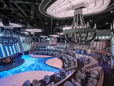 Panorama Lounge der MSC World Asia. Bühne mit Instrumenten, gestufte Sitzbereiche, blauer Wasser-Effekt-Boden und Meerblick. Kronleuchter spenden Licht. Keine Personen.