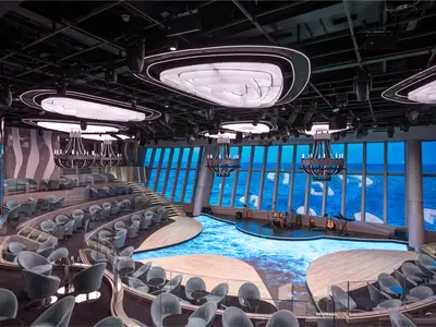 Moderne Panorama Lounge an Bord der MSC World Asia. Eine Buehne mit Instrumenten, gestufte Sitzbereiche und Meerblick. Futuristische Deckenleuchten und ein leuchtender Boden.