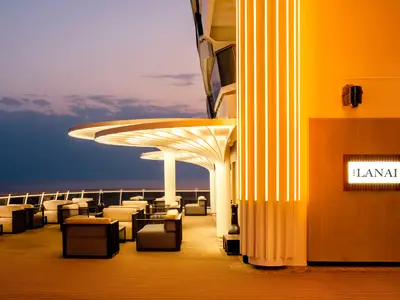 Dunkelblaues Meer und Dämmerungshimmel über dem beleuchteten Außenbereich The Lanai an Bord der MSC World Asia. Komfortable Sitzgruppen unter geschwungenen, leuchtenden Dächern und einer gelb-orange beleuchteten Wand.